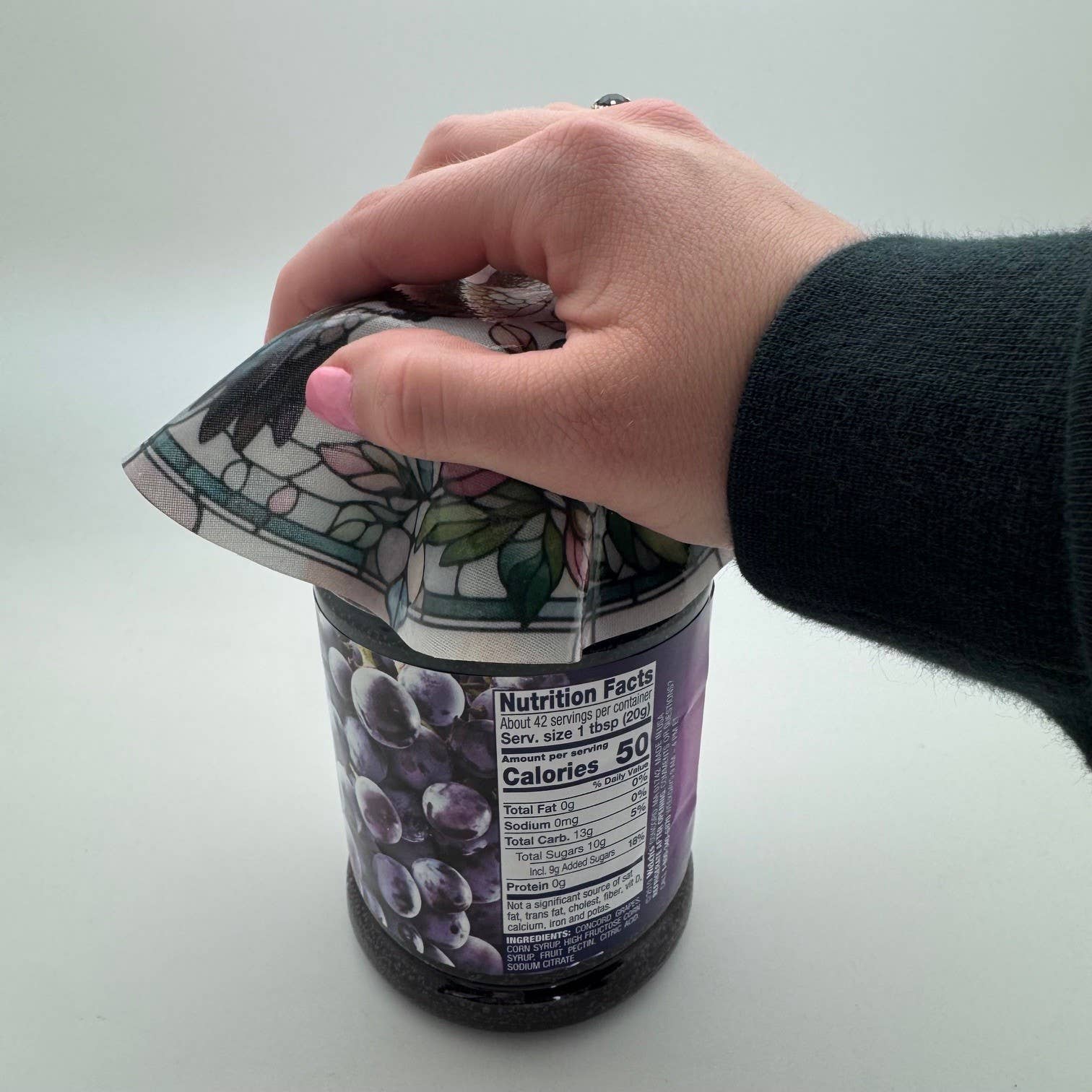 Hand using the Hummingbird Stained Glass Silicone Jar Opener by Andréas to open a grape jelly jar, demonstrating the non-slip silicone grip in action — stained glass hummingbird design visible on the flexible disc