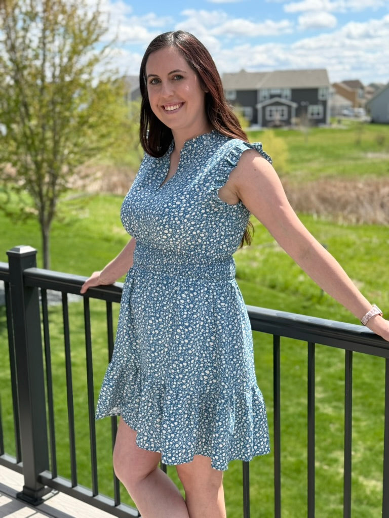 Woman in a blue floral dress standing on a balcony with a green landscape and houses in the background.