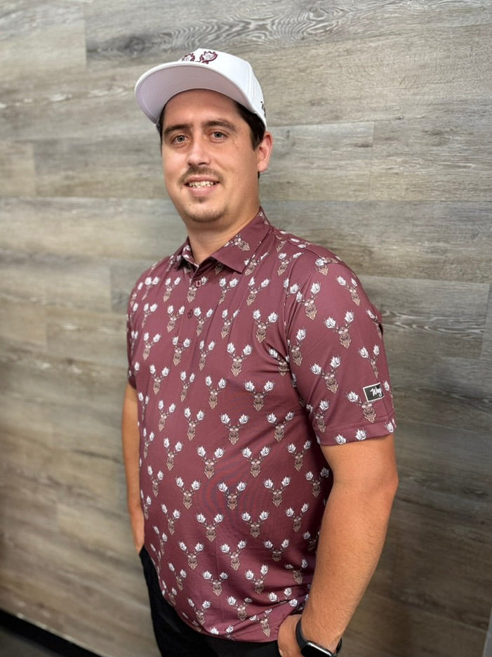 A man wearing a Waggle Rack Star burgundy short-sleeve polo shirt with a moose pattern, paired with a white baseball cap, standing against a gray wood-paneled wall.