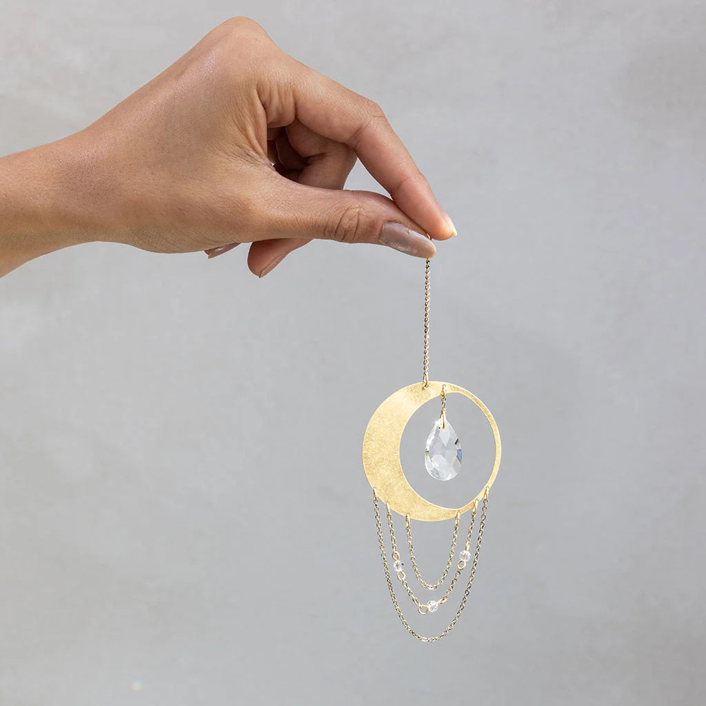 Hand holding Scout Curated Wears Crescent Moon Balance mini crystal suncatcher featuring gold moon design with crystal prism and rainbow reflections
