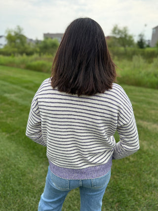 A person viewed from behind wearing a cream and purple horizontally striped knit sweater with a purple ribbed hem, standing on a grassy outdoor lawn.