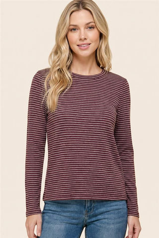 A woman modeling a long-sleeve crewneck top with thin horizontal burgundy and white stripes, paired with blue jeans, against a solid beige background.
