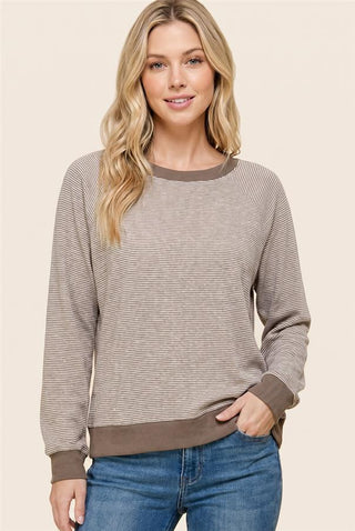 A woman wearing a long-sleeve crewneck sweater with fine horizontal stripes in taupe and brown, paired with blue jeans, against a solid light beige background.