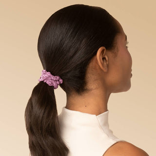A woman shown from the back and side profile wearing a textured lavender scrunchie ponytail against a solid beige background.