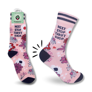 A pair of pink crew socks with a floral print and the text 'NEXT STOP THRIFT SHOP' in navy blue. One sock is shown hanging from its green and white Funatic retail tag, while the other is displayed flat.