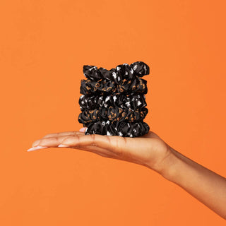 A hand holding a stack of black satin scrunchies with a small ghost or pumpkin pattern, set against a solid orange background.