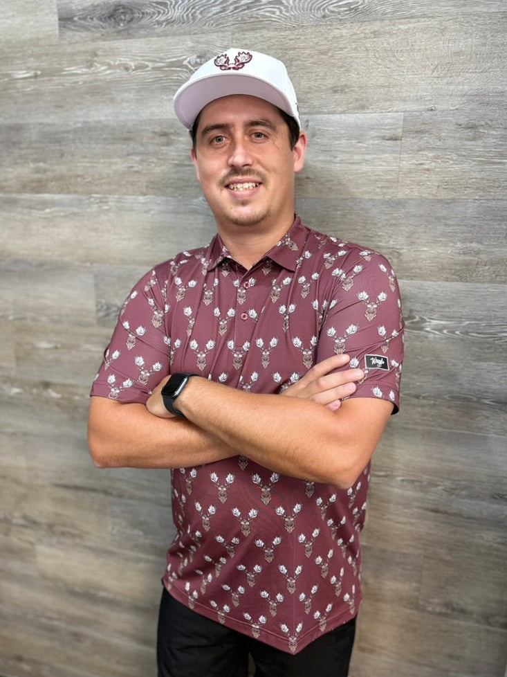 A man wearing a Waggle Rack Star burgundy short-sleeve polo shirt with a moose pattern, a white baseball cap, and black pants, standing with arms crossed against a gray wood-paneled wall.