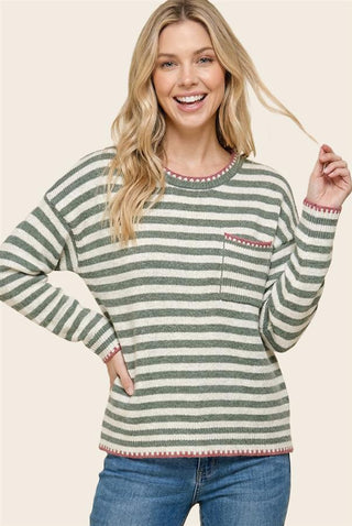 A woman wearing a green and white striped long-sleeve knit sweater with a chest pocket and pinkish-red trim, paired with blue jeans, against a solid light beige background.