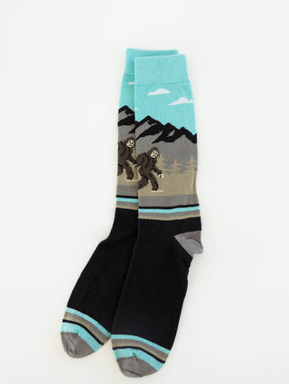 A pair of 1902 crew socks with a graphic design of Bigfoot walking in front of mountains. The upper portion features a light blue sky and mountain landscape, while the lower portion is black with gray and light blue striped accents on the toes and heels.