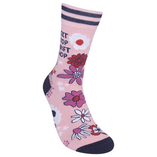 A single pink crew sock with a floral pattern, featuring dark navy blue stripes at the ribbed cuff, a navy heel, toe, and sole, and text reading 'NEXT STOP TRIFT SHOP'.