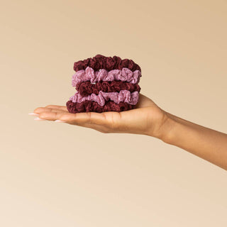 A hand holding a stack of four fabric scrunchies in shades of burgundy and dusty pink against a solid beige background.