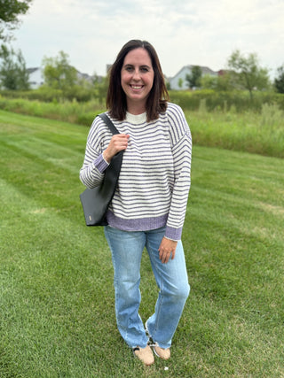 A woman stands on a grassy lawn wearing a cream and purple striped knit sweater with a purple hem and cuffs, light blue jeans, and beige shoes, while carrying a black shoulder bag.