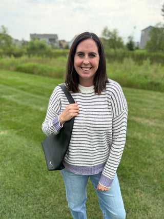 A woman standing outdoors on a grassy lawn wearing a cream and purple striped knit sweater, Risen blue jeans, and carrying a black hobo shoulder bag.