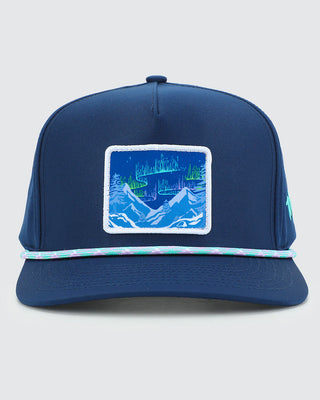 A navy blue snapback cap featuring a rectangular embroidered patch on the front with a blue aurora landscape and a thin pastel braided trim across the brim.