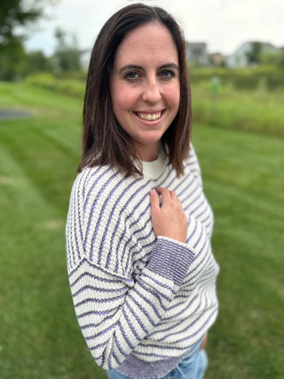 A woman smiling outdoors on a grassy lawn, wearing a knit sweater with horizontal cream and purple stripes.