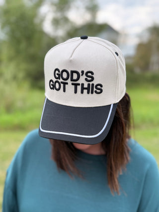 A person outdoors wearing a baseball cap with the text 'GOD'S GOT THIS' in black on an off-white crown and a black brim.