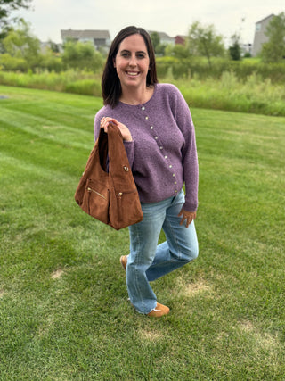 A woman stands on a green lawn wearing a knit cardigan, Risen jeans, and tan shoes, while holding a brown Heidi faux suede shoulder bag.