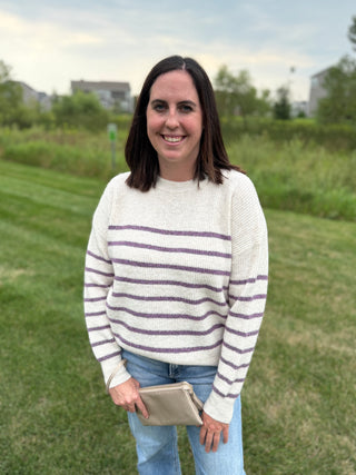 A woman standing outdoors on a grassy lawn wearing a cream knit sweater with purple horizontal stripes, blue jeans, and holding a beige clutch.