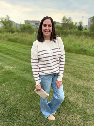 A woman standing on a grassy lawn wearing a cream knit sweater with purple horizontal stripes, blue jeans, and beige shoes, holding a small beige clutch.