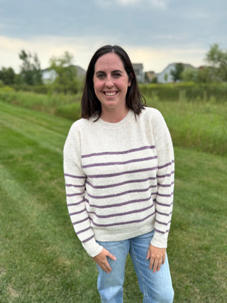 A woman standing outdoors on a grassy lawn wearing a cream knit sweater with purple horizontal stripes and light blue jeans.