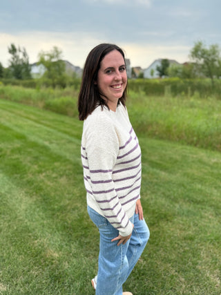 A woman standing on a grassy lawn wearing a cream knit sweater with purple horizontal stripes and light blue jeans.