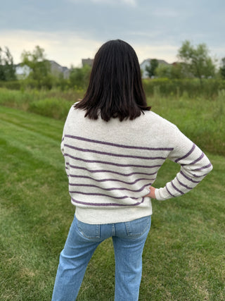 A woman viewed from behind wearing a cream knit sweater with dark purple horizontal stripes and light blue jeans, standing on a grassy lawn.