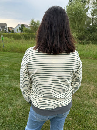 A person viewed from behind wearing a cream-colored long-sleeve top with thin green horizontal stripes and light blue jeans, standing on a grassy lawn outdoors.