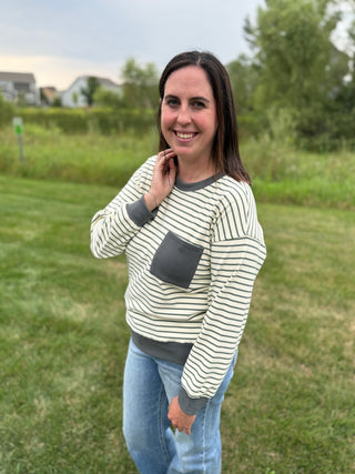 A woman standing outdoors on a grassy lawn wearing a cream and dark gray striped long-sleeve top with a solid dark gray chest pocket and matching dark gray cuffs and hem, paired with blue jeans.