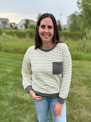 A woman standing outdoors on a grassy lawn wearing a long-sleeve crewneck sweatshirt with thin dark green horizontal stripes on an off-white base, featuring a solid dark gray pocket and ribbed collar, cuffs, and hem.