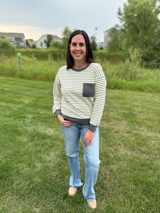 A woman standing on a grassy lawn wearing a cream and dark green striped long-sleeve top with a dark gray pocket, paired with light blue jeans and tan shoes.