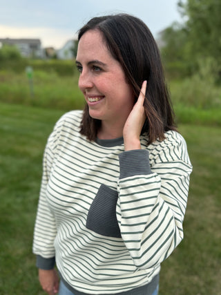 A woman wearing a cream and dark green striped long-sleeve top with dark green ribbed cuffs and a pocket, standing outdoors in a grassy field.