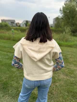 Back view showing the hood and ribbed hem, layered over a blue floral long-sleeve top with jeans, outdoors.