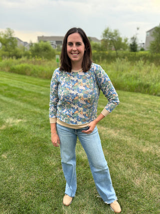 A woman standing on a grassy lawn wearing a long-sleeve crewneck long-sleeve top with a blue, green, and yellow floral pattern, paired with light blue jeans and beige shoes.
