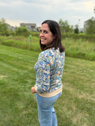 A woman wearing a blue and beige floral patterned long-sleeve top with light blue jeans, looking back over her shoulder.
