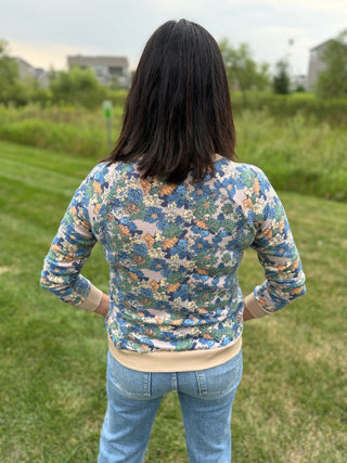 A woman viewed from behind wearing a long-sleeve crewneck sweatshirt with a blue, green, and beige floral pattern, paired with light blue jeans.