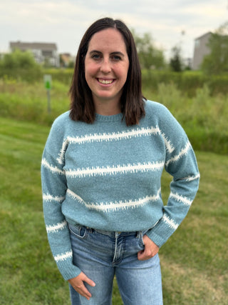 A woman standing outdoors on a grassy lawn wearing a light blue knit sweater with white horizontal textured stripes and blue jeans.