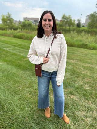 A woman standing on a grassy lawn wearing a cream and taupe striped long-sleeve hoodie, blue wide-leg jeans, and tan clogs, with a small plum crossbody bag.