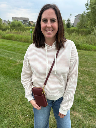 A woman standing outdoors on a grassy lawn wearing a cream and taupe horizontally striped hoodie, blue jeans, and carrying a small plum crossbody phone pouch with gold hardware.