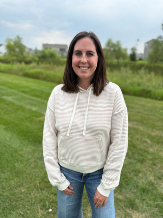 A woman standing outdoors on a grassy lawn wearing a cream and taupe horizontally striped pullover hoodie with cream drawstrings and blue jeans.