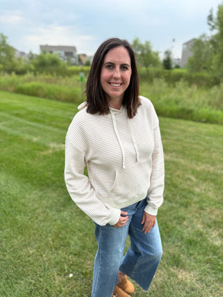 A woman standing outdoors on a grassy lawn wearing a cream and taupe colored, horizontally textured pullover hoodie with drawstrings and blue jeans.