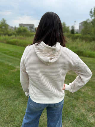 A person viewed from behind wearing a cream and taupe colored, horizontally ribbed knit hoodie and blue jeans, standing on a grassy lawn with trees and buildings in the background.