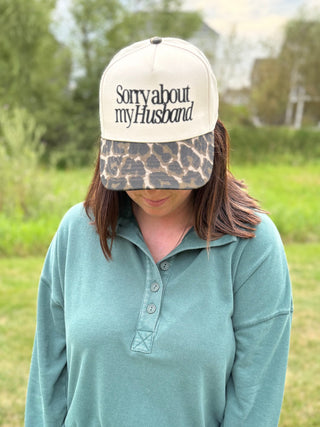 A woman wearing a sage green long-sleeve shirt and a cream trucker hat with a leopard-print brim. The hat features the text 'Sorry about my Husband' in black.