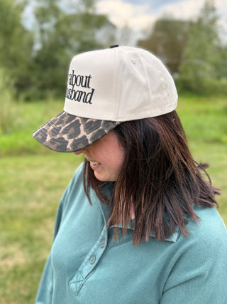 A woman wearing a baseball cap with a cream crown and a leopard-print visor, featuring the embroidered text  'sorry about husband' on the front.