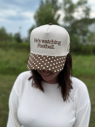 A woman wearing a cream and brown trucker hat with the text 'He's watching Football.' and a polka-dot visor.