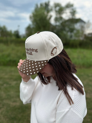 A woman outdoors wearing a cream baseball cap with brown polka-dot visor trim and brown embroidered text reading 'he's watching football.' on the front, paired with a white top.