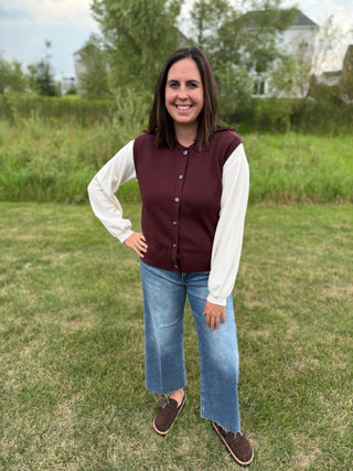 A woman standing outdoors wearing a plum button-up vest and a white long sleeve top, blue wide-leg jeans, and brown slip-on shoes.