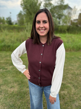 A woman standing outdoors wearing a plum sweater vest and white top with blue jeans.