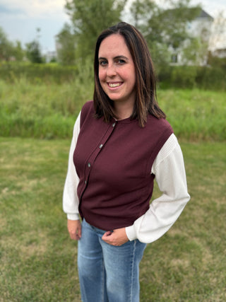 A woman standing outdoors wearing a plum ribbed knit cardigan vest over a white long-sleeve top and blue jeans.