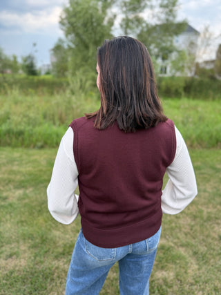 A woman viewed from behind wearing a plum ribbed knit sweater vest over a white long-sleeve shirt and blue jeans.