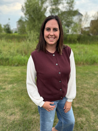 A woman standing outdoors wearing a sleeveless plum button-up cardigan with white long sleeves and blue jeans.